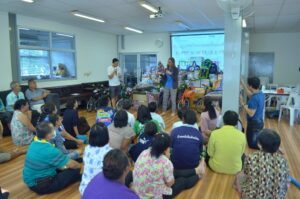 Vera Hofbauer und ein Kollege halten einen Vortrag in Thailand vor einigen Menschen, die in einem Klassenzimmer am Boden sitzen