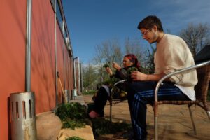 Ein junger Mann und eine ältere Frau sitzen im Freien auf Sesseln. Die halten beide ein Büschel Grün in der Hand.