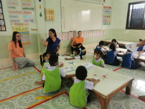 Volunteers (4) Zwei Freiwillige und eine Lehrerin sitzen in einer Schulklasse und unterrichten Kinder.