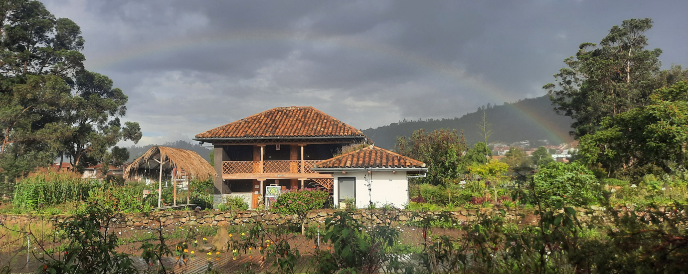 Ein Haus mit braunem Dach, rings herum grüne Bäume, ein Regenbogen.