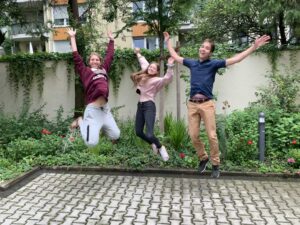 Drei junge Menschen springen mit erhobenen Armen in die Luft. Im Hintergrund ein Innenhof.