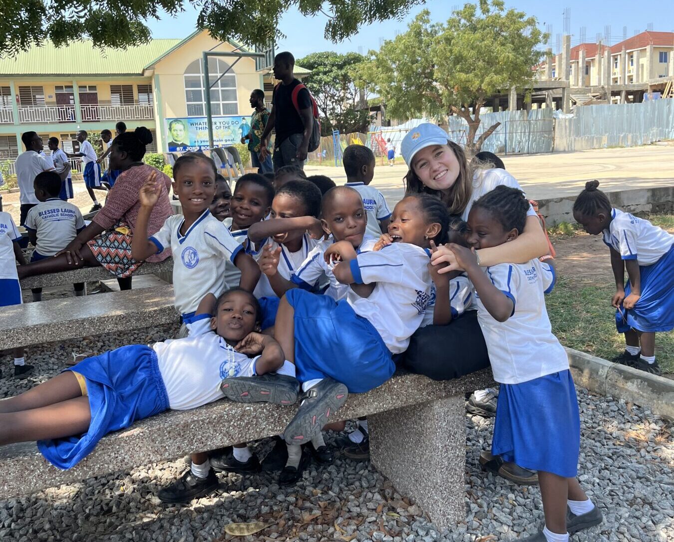 Eine junge Frau sitzt umringt von vielen Kindern an einem Tisch. Sie haben unterschiedliche Hautfarben. Im Hintergrund sind weitere Personen und ein Baum zu sehen.