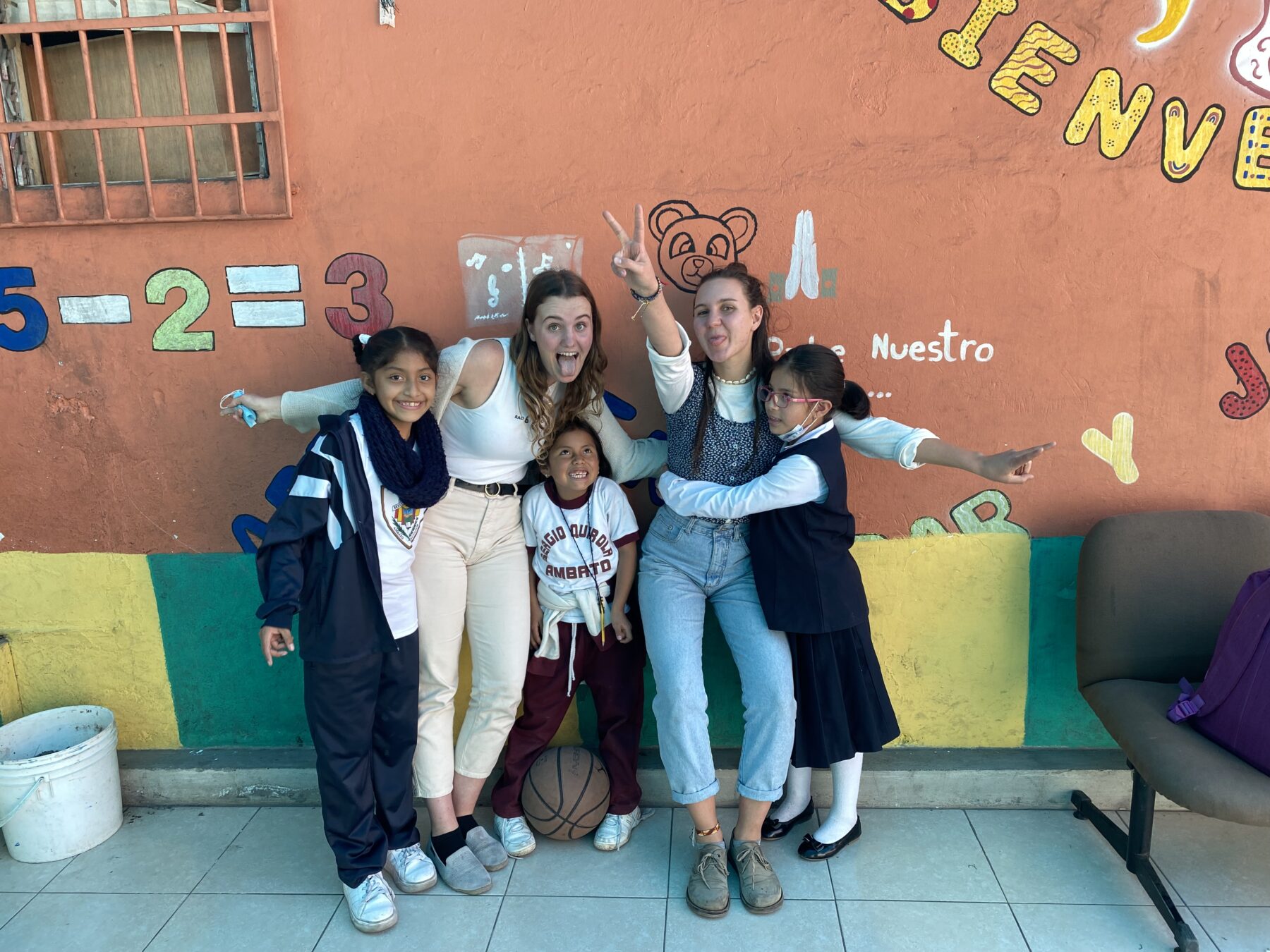 Zwei junge Frauen und drei Kinder stehen nebeneinander vor einer orangenen Wand. Sie lachen und schneiden Grimassen in die Kamera.