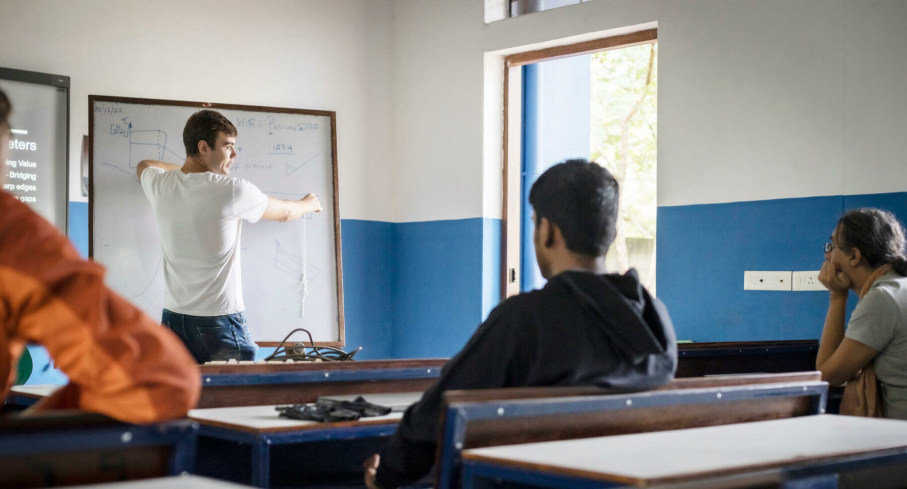 © Aparna Jayakumar/ WeltWegWeiser/ Fairpicture Ein junger Mann steht an der Tafel, mit dem Rücken zur Kamera. Hinter ihm sitzen einige Schüler:innen an Schultischen.