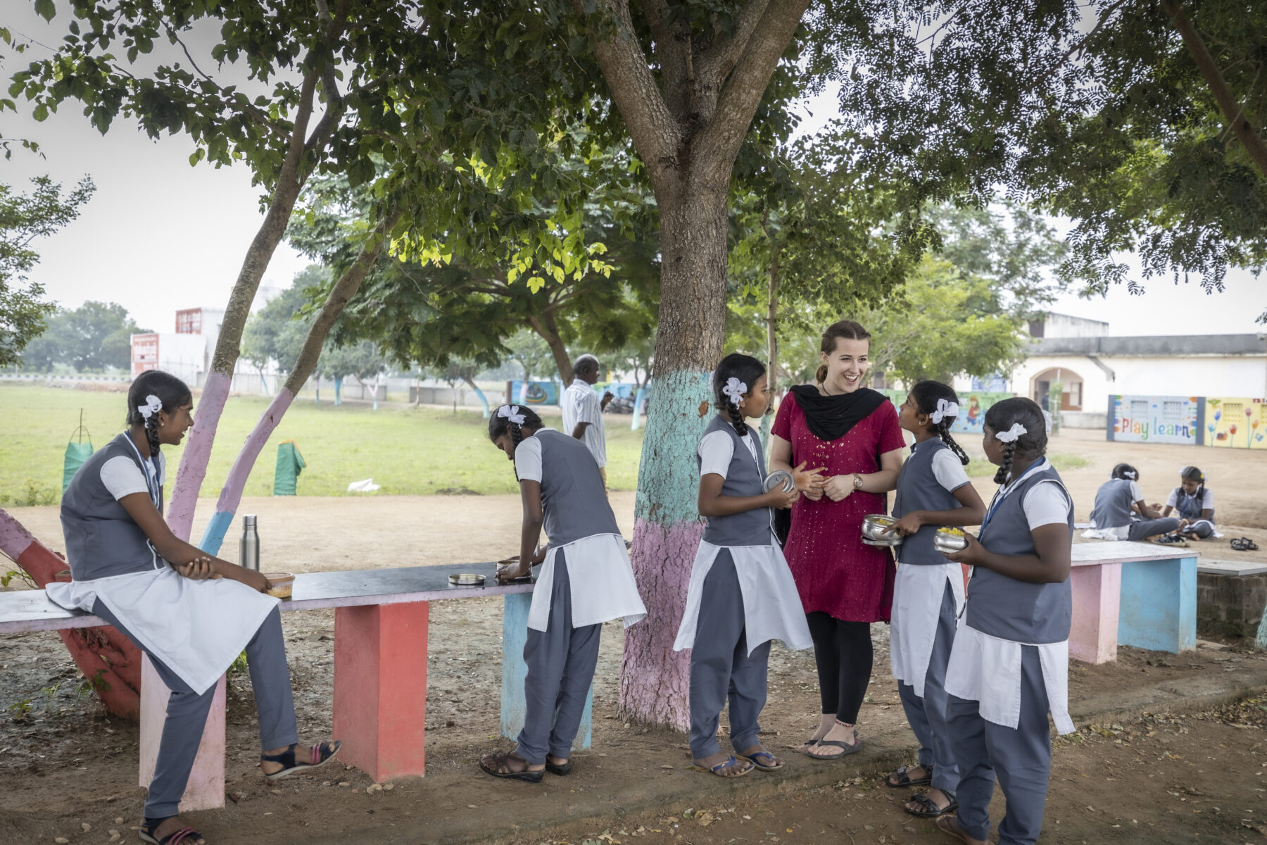 © Aparna Jayakumar/ WeltWegWeiser/ Fairpicture eine junge Frau und ein paar Mädchen in Schuluniformen stehen unter einem Baum.