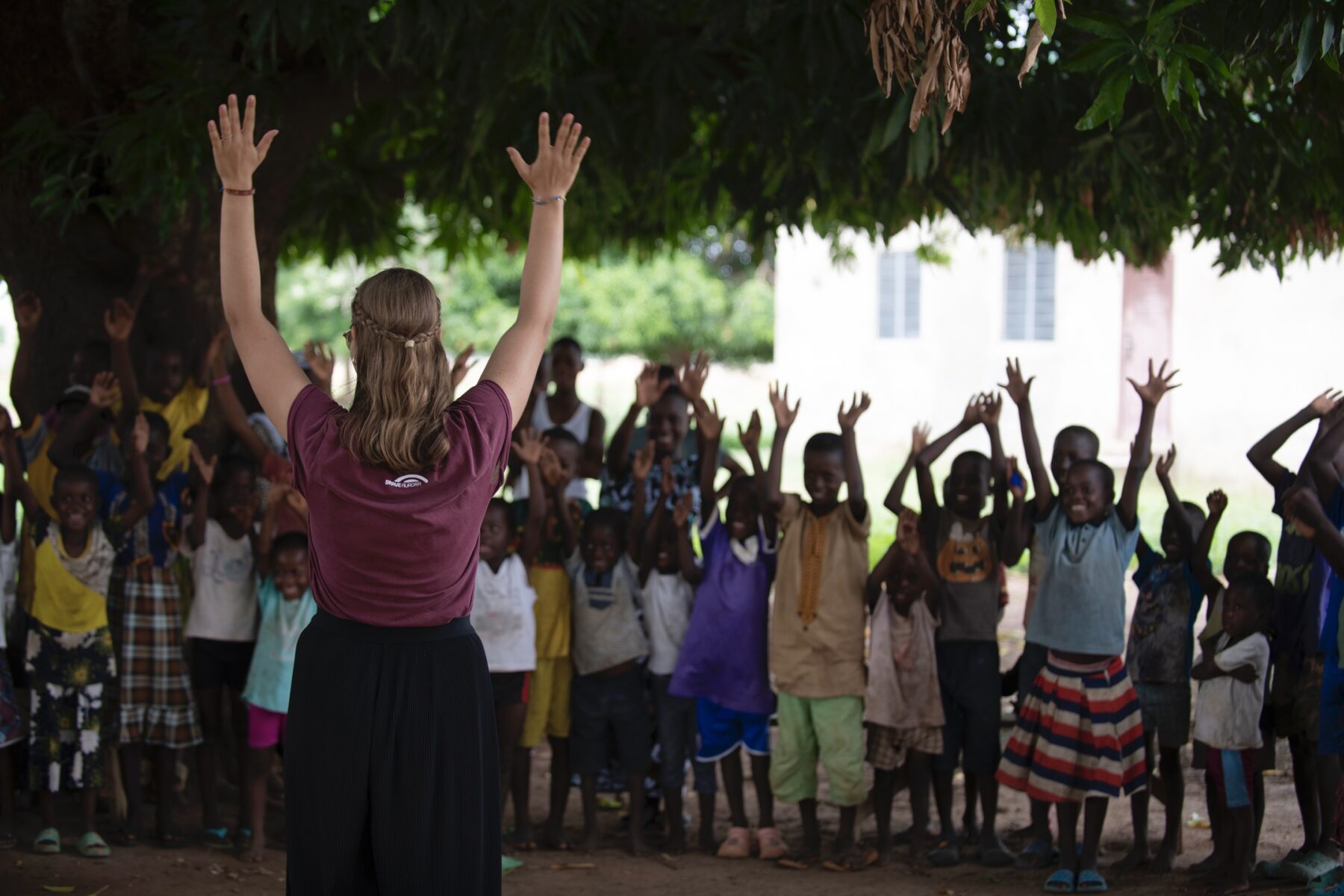 c Victoria Hausleitner x Braveaurora Eine junge Frau steht mit dem Rücken zur Kamera. Sie hält die Hände in die Höhe. Gegenüber steht eine Gruppe von Kindern, ebenfalls mit den Händen in der Höhe.
