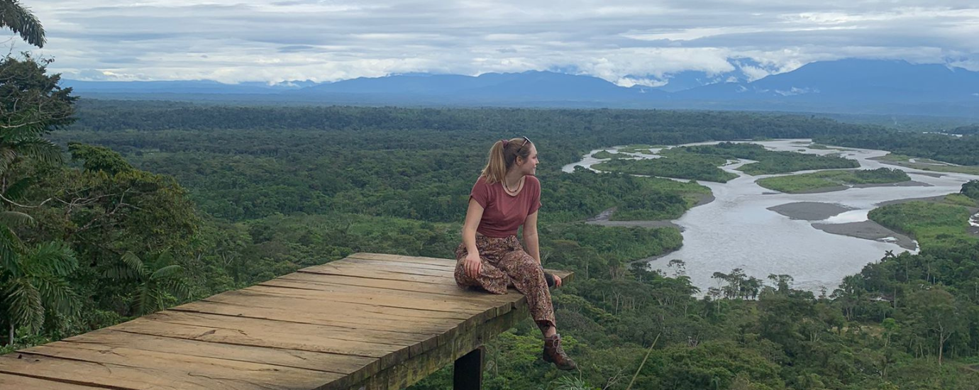 Eine junge Frau sitzt auf einem Steg hoch in der Luft und schaut auf einen Wald mit einem großen Fluss.