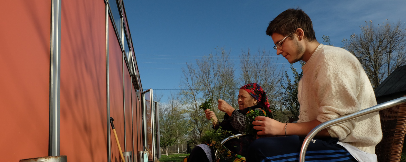 Ein junger Mann und eine ältere Frau sitzen im Freien nebeneinander und halten Kräuter in der Hand.