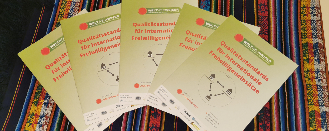 c WeltWegWeiser Mehrere Hefte liegen auf einem bunten Tischtuch. Darauf steht "Qualitätsstandards für internationale Freiwilligeneinsätze".