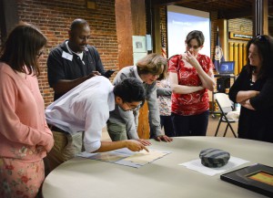 Eine Gruppe von Erwachsenen steht rund um einen Tisch, ein Mann zeigt auf ein Dokument, das am Tisch liegt