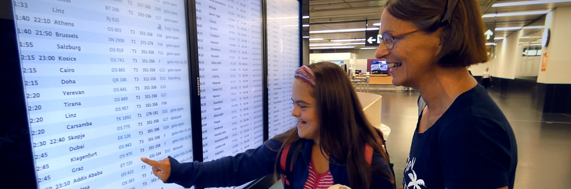 Eine junge Frau und ihre Mutter stehen vor einer Anzeigetafel am Flughafen