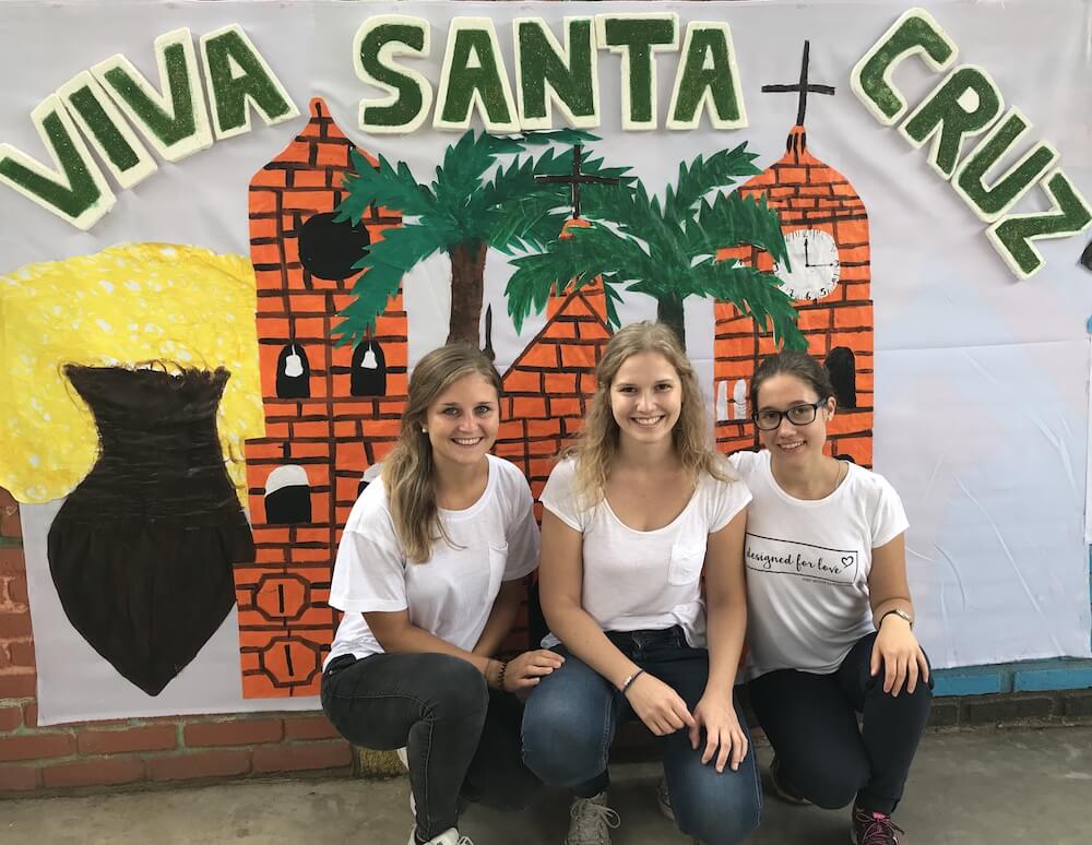 Drei weibliche Freiwillige hocken vor einem Wandbild, auf der Viva Santa Cruz geschrieben steht