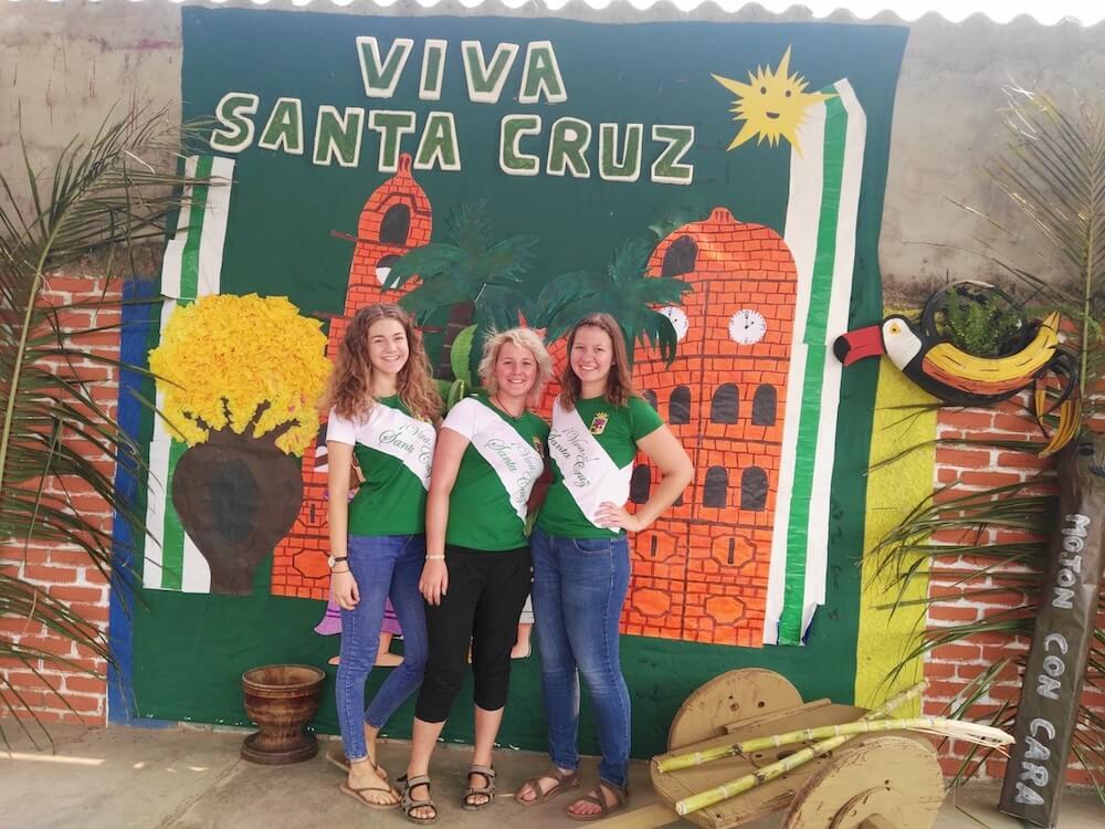Drei weibliche Freiwillige stehen vor einem Wandbild mit der Schrift Viva Santa Cruz, sie tragen grüne T-Shirts mit einer Schärpe