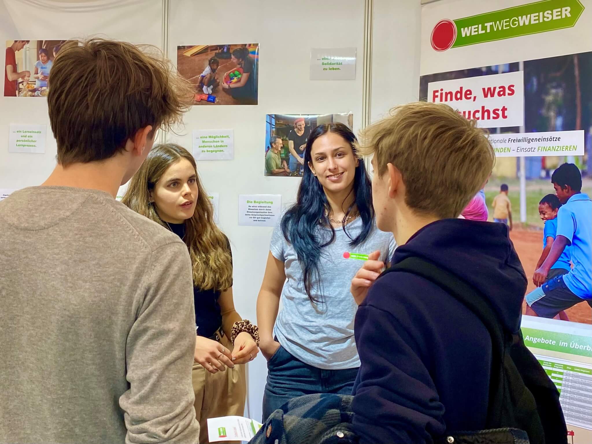 Multiplikatorinnen beraten bei einem Messestand von WeltWegWeiser zwei interessierte Jugendliche über einen Freiwilligeneinsatz