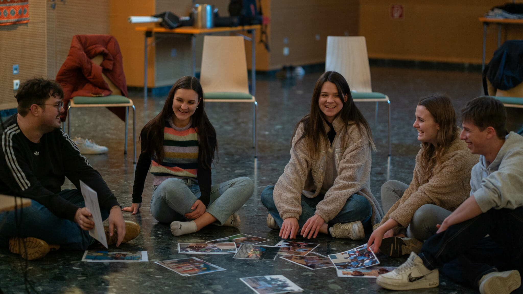 Mehrere junge Personen sitzen im Kreis am Boden, in der Mitte liegen Fotos, sie lachen